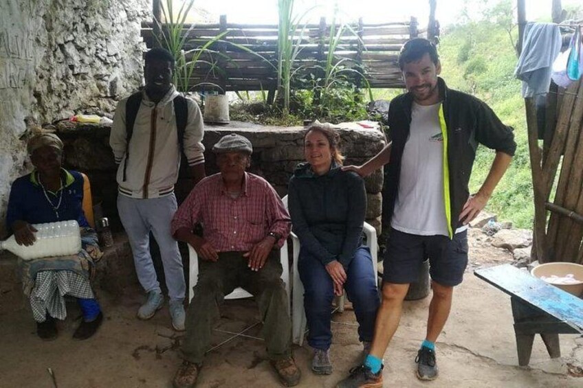 A group of people in an outdoor setting with a rustic background, including a stone wall and wooden structures. 2