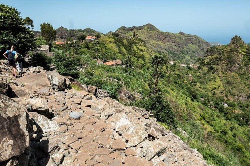 A rocky hiking trail with hikers, lush green hills, and distant houses. 5