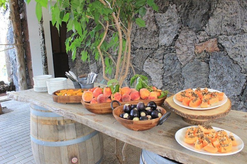 A wooden table with various fruits and vegetables, including peaches, black olives, and carrots, set against a stone wall and a tree. 4