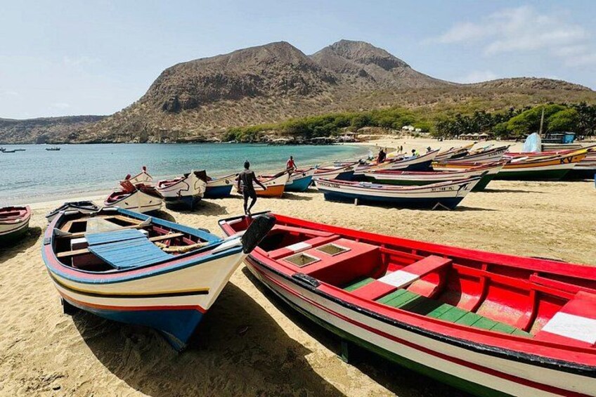 A beach with several colorful boats lined up, a person standing, and a mountain in the background. 3