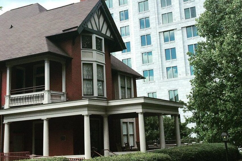 A Victorian-style house with a balcony and a large porch, surrounded by a well-maintained garden and a modern building in the background. 1