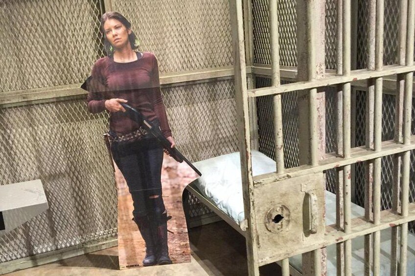 A person standing in a cell with bars, holding a gun, and a bed visible in the background. 4