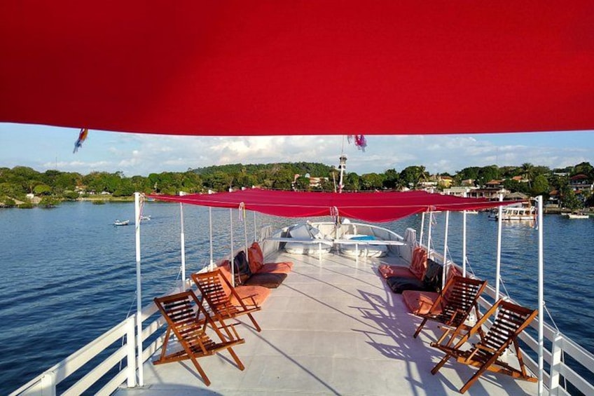A boat deck with wooden chairs and a red canopy, overlooking a calm body of water with boats and a shoreline in the background. 2