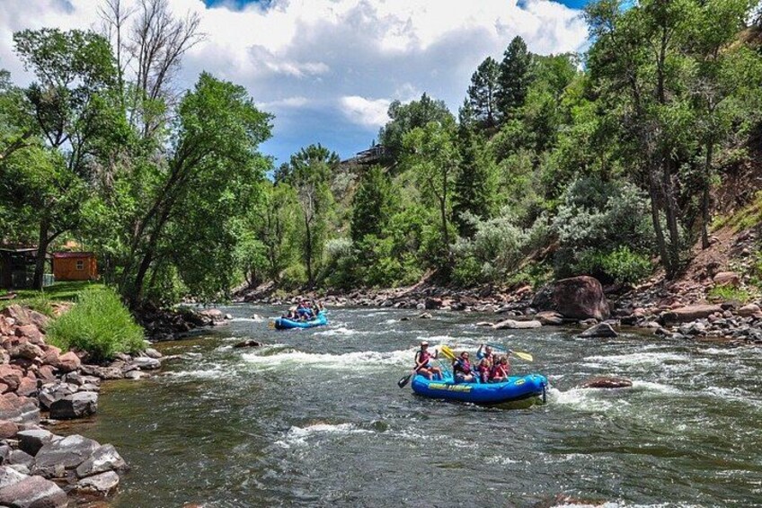 A river with two blue inflatable rafts with people, surrounded by trees and rocks. 2