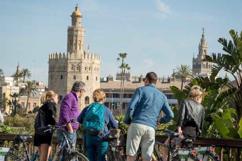 A group of people standing on a bridge with bicycles, overlooking a historic tower and other buildings. 3
