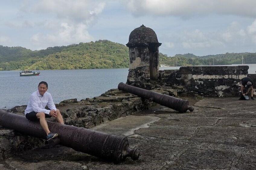 A historical fort with cannons, a person sitting on a cannon, and a boat on the water. 2