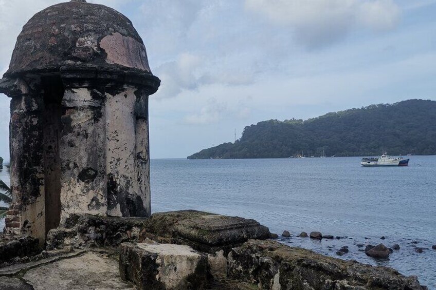 A weathered lighthouse by the sea, a boat in the distance, and a forested hill. 1