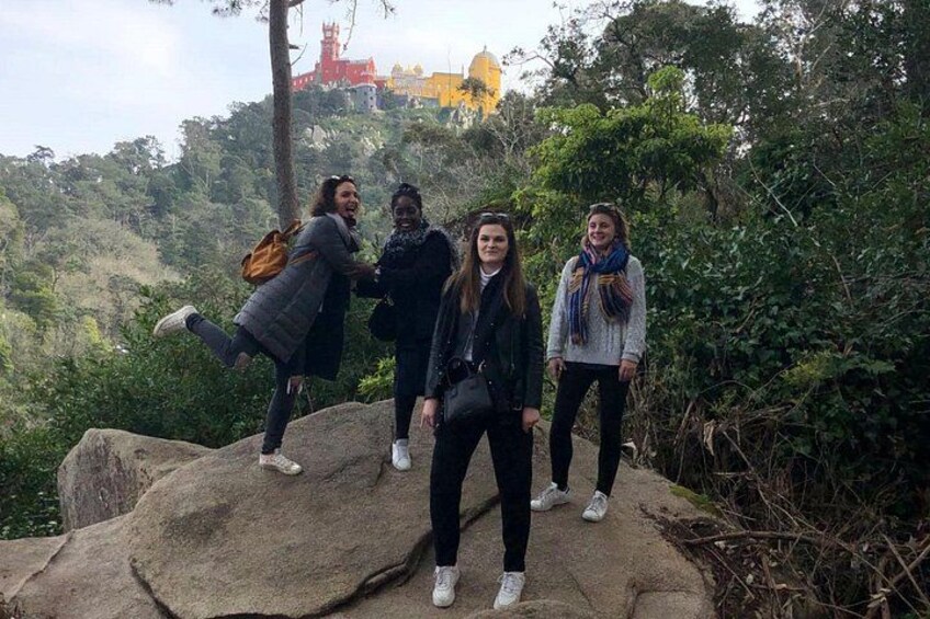 Four people standing on a rock with a forested hill and a red building in the background. 4