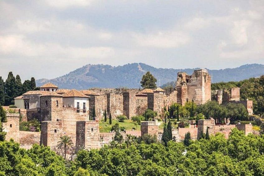 A historical site with ruins, including a large tower and several buildings, surrounded by greenery and mountains in the background. 2