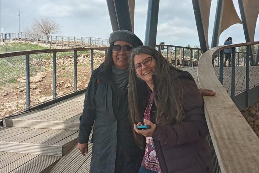 Two people standing on a wooden boardwalk with metal railings, overlooking a rocky landscape. 2