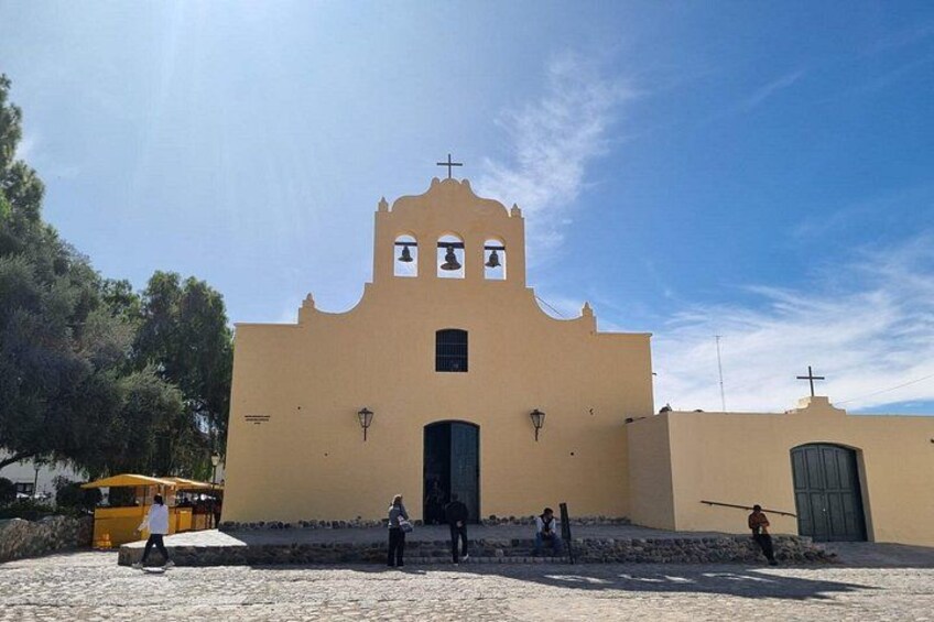Una iglesia con un campanario, una puerta grande y una ventana pequeña. Hay gente parada frente al edificio. 5