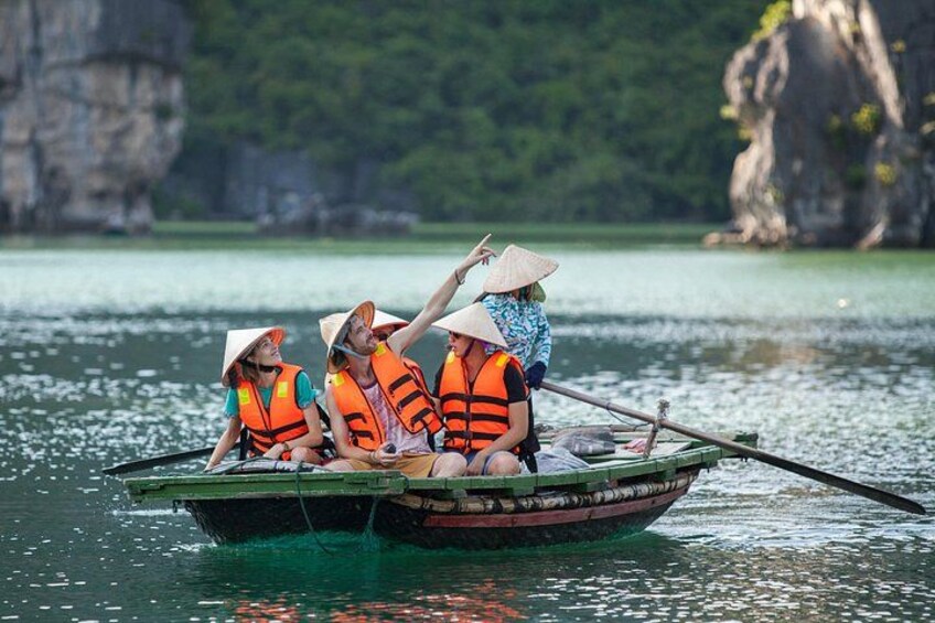 A group of people in a boat wearing life jackets and conical hats, paddling on a calm body of water with rocky cliffs in the background. 2