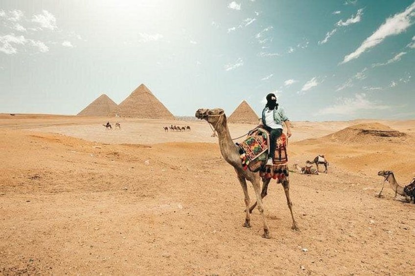 A camel with a rider in a desert landscape with pyramids in the background. 3