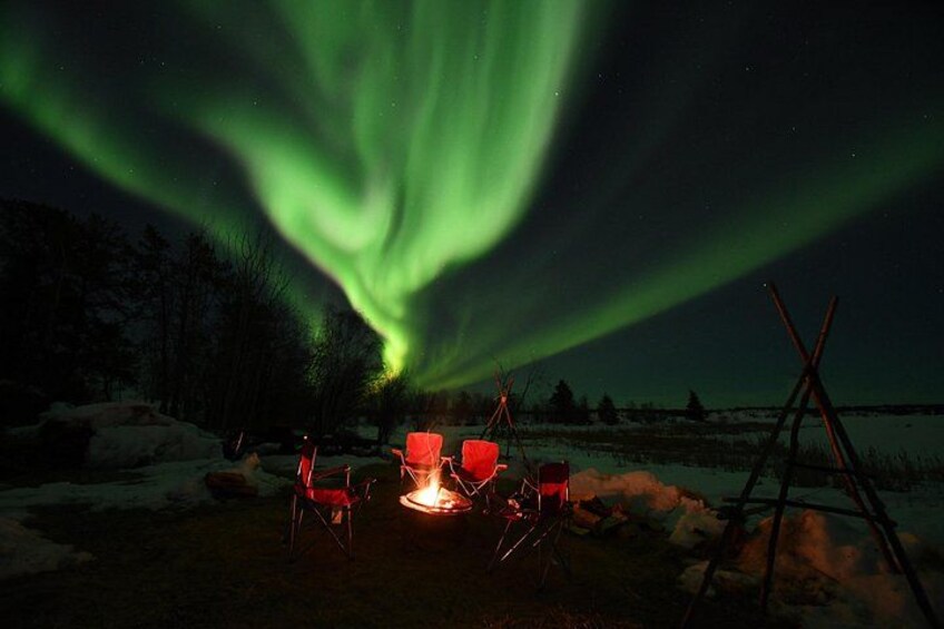 Una fogata con sillas a su alrededor, un paisaje nevado y las auroras boreales en el cielo. 4