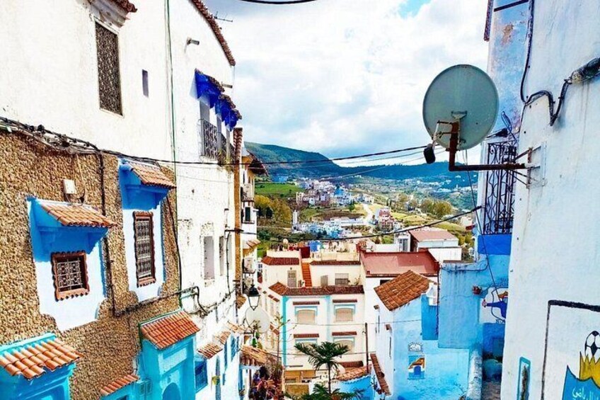A narrow street with buildings painted in blue and white, a satellite dish, and a view of a town in the distance. 5