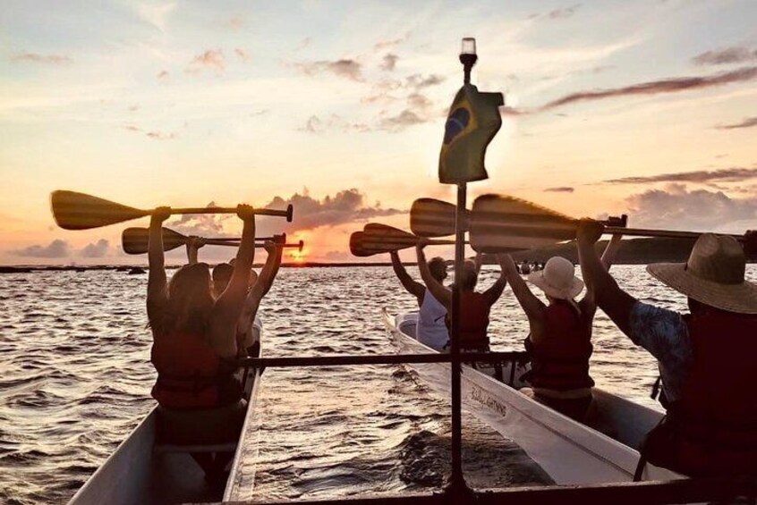 Personas en un bote remando con remos, una bandera y un atardecer al fondo. 1