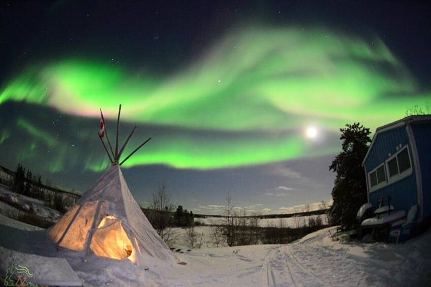 Paisaje nevado con un tipi iluminado y una cabaña azul bajo un cielo nocturno con auroras boreales verdes. 3