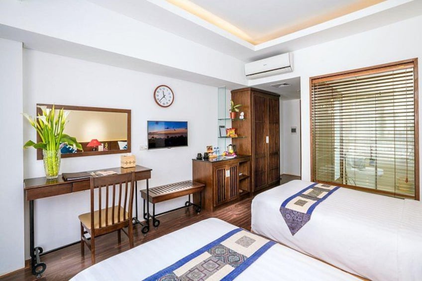 A hotel room with two beds, a desk, a chair, a TV, a clock, and a window with blinds. 3