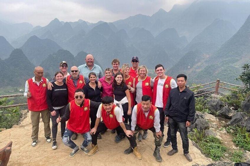 A group of people in red vests posing on a mountain ridge with a view of distant mountains. 2