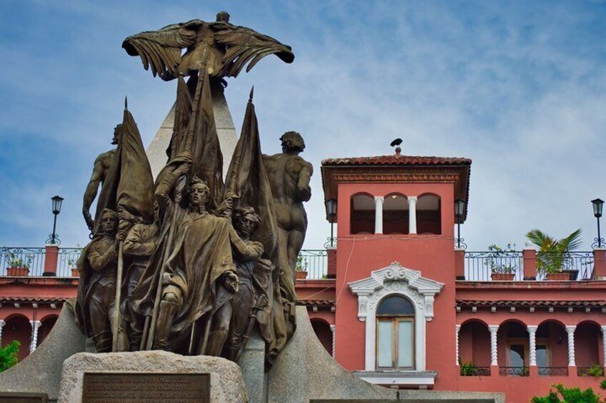 A historical monument with a central figure holding a staff, surrounded by figures in dynamic poses, set against a backdrop of a traditional building with arched windows and a red facade. 4
