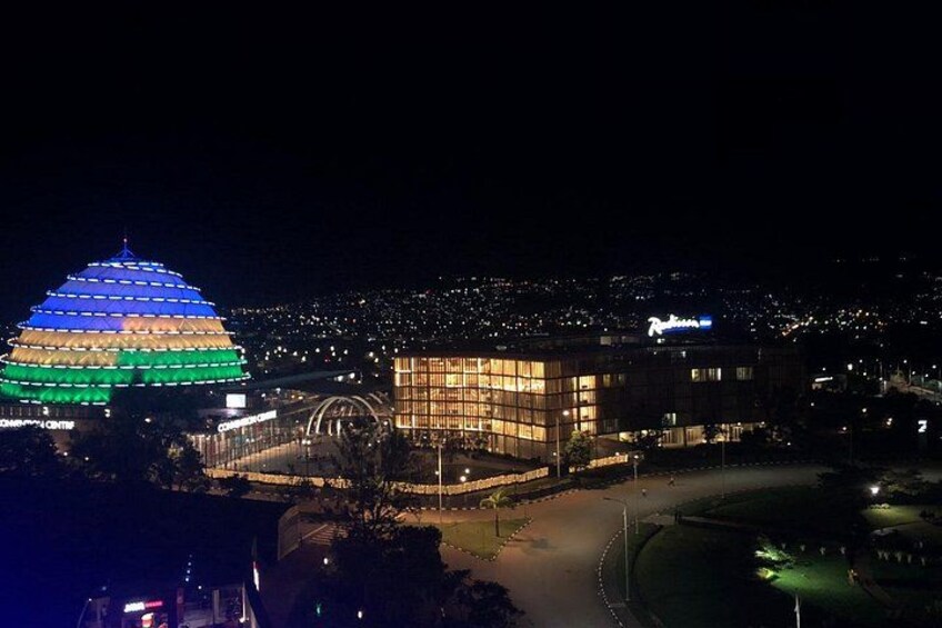 Una estructura en forma de cúpula, muy iluminada, con un exterior de colores variados, un edificio moderno con ventanas iluminadas y un horizonte urbano lejano con luces. 1