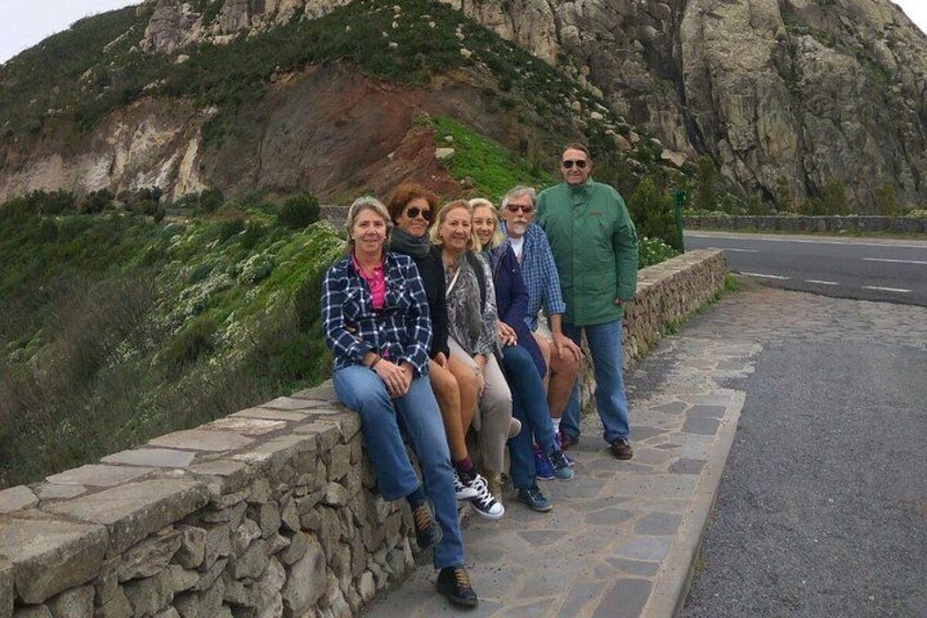 A group of people sitting on a stone wall by a road with a mountainous landscape in the background. 1