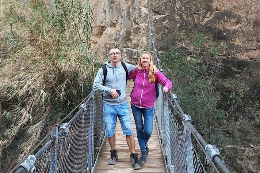 Two people standing on a suspension bridge with metal railings and wooden planks, surrounded by dry vegetation and rocky cliffs. 3