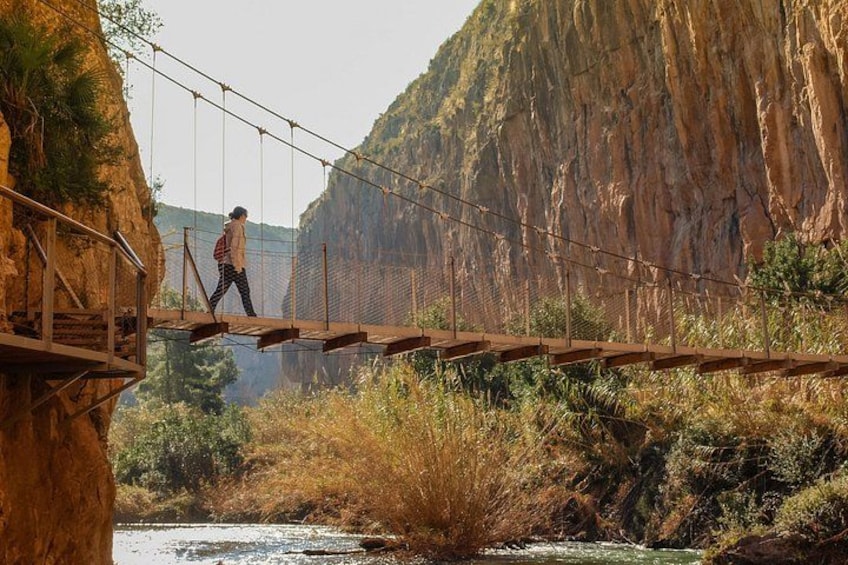 A person walking on a suspension bridge over a river with cliffs in the background. 1