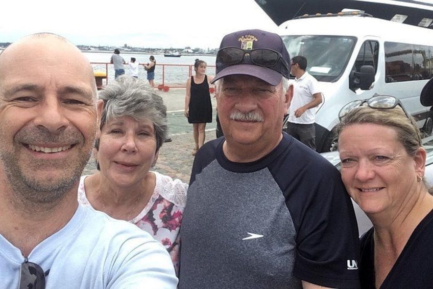 Un grupo de personas posando para una foto cerca de un cuerpo de agua, con un bote y un vehículo blanco al fondo. 1