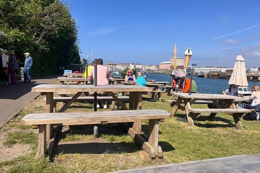 Tavoli da picnic all'aperto in riva all'acqua, con persone sedute e che passeggiano. 5