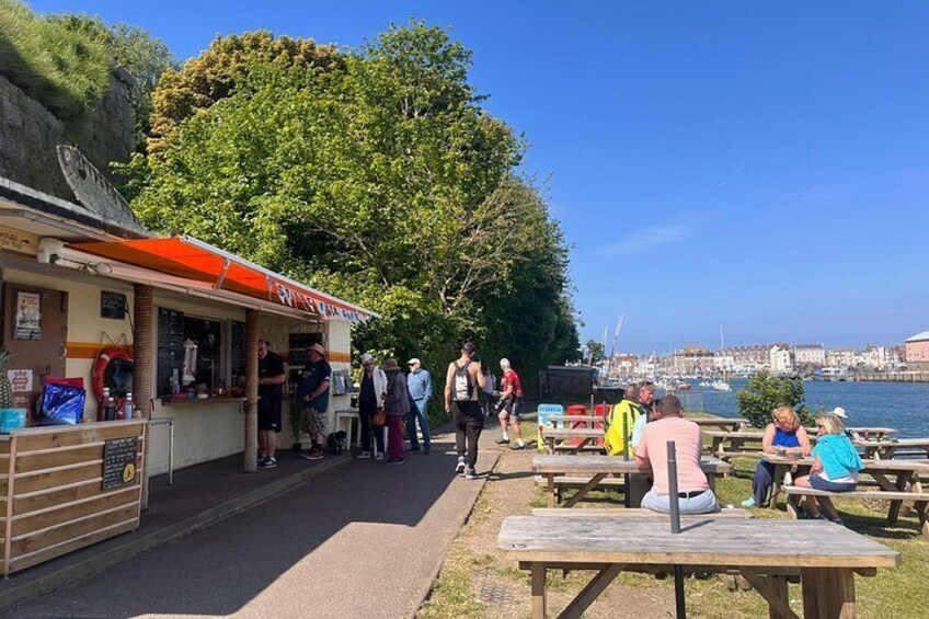 Area picnic lungo il fiume con persone che cenano, un chiosco gastronomico e vista sulla città dall'altra parte dell'acqua. 2