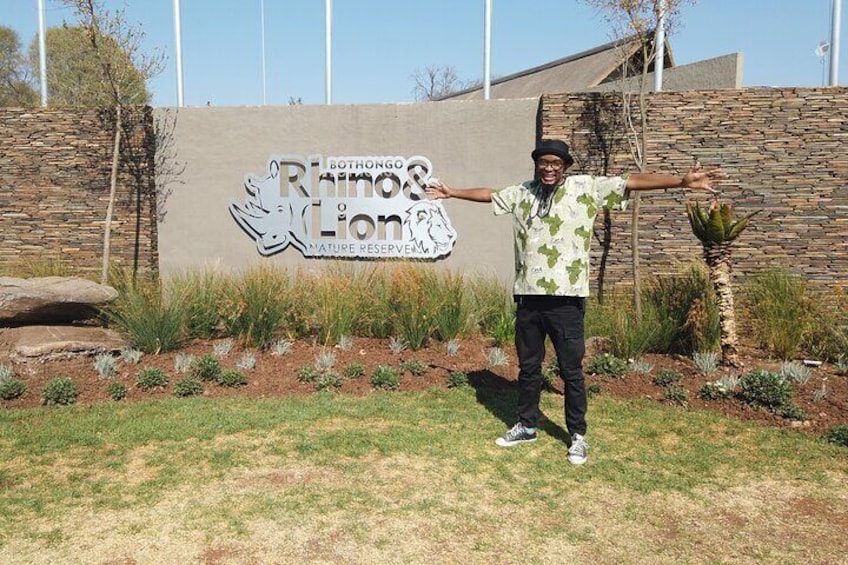 A person standing in front of a sign with a lion graphic and the text 'Rhinos & Lions Nature Reserve.' 2