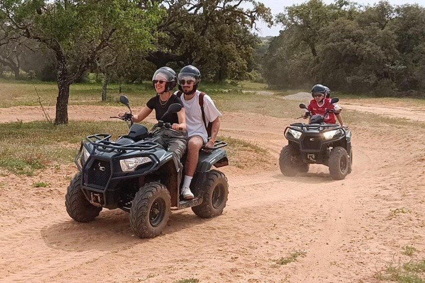 Two people riding an ATV on a dirt path with trees in the background. 2