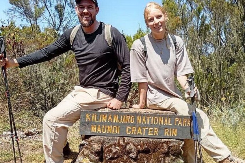 Dos personas sentadas sobre una roca con un letrero que dice 'Kilimanjaro National Park Maundi Crater Rim'. 2