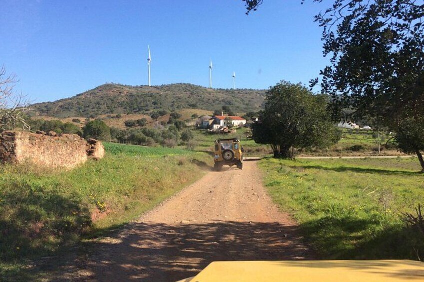 A dirt road with a yellow vehicle, wind turbines in the background, and a hilly landscape. 3