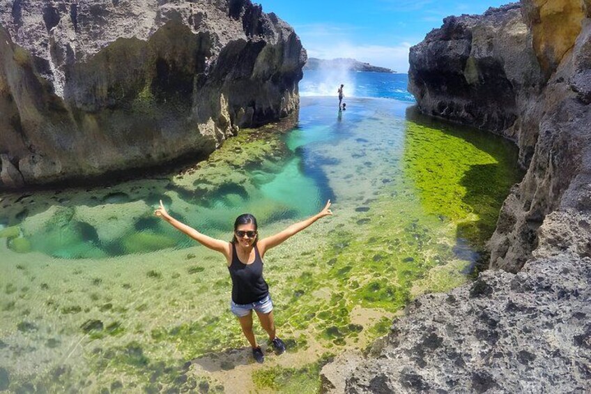 A person standing on a rocky outcrop with arms outstretched, overlooking a clear blue sea with visible underwater rocks and a distant person in the water. 5