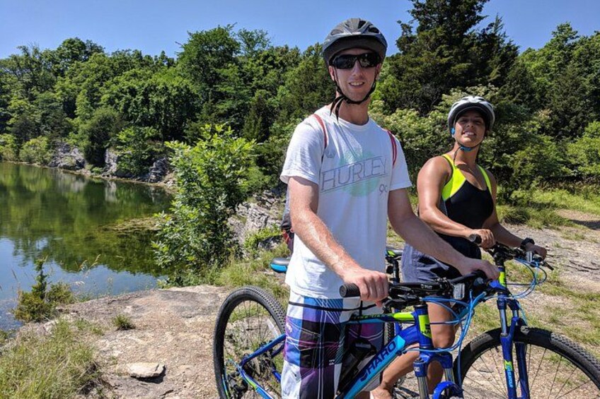 Two people on bicycles near a body of water with trees in the background. 4