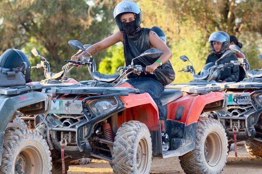A person on a red ATV with a black bag, another person in the background on a similar ATV, and a natural outdoor setting. 1
