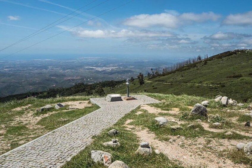 A paved pathway leading to a viewpoint with a stone monument, surrounded by grassy terrain and scattered rocks. 1