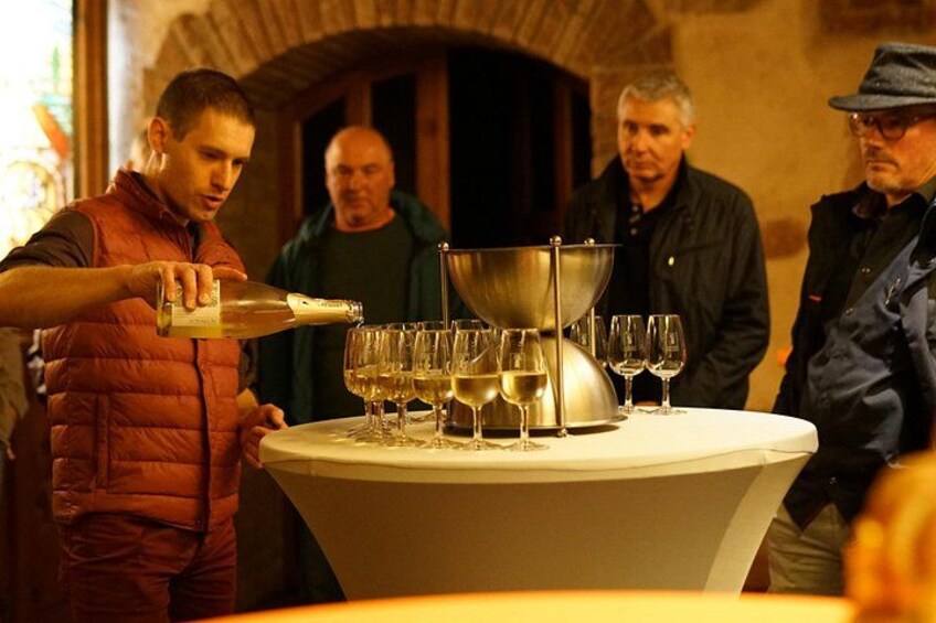 A person pouring a liquid into a large metal container on a table surrounded by wine glasses. 1