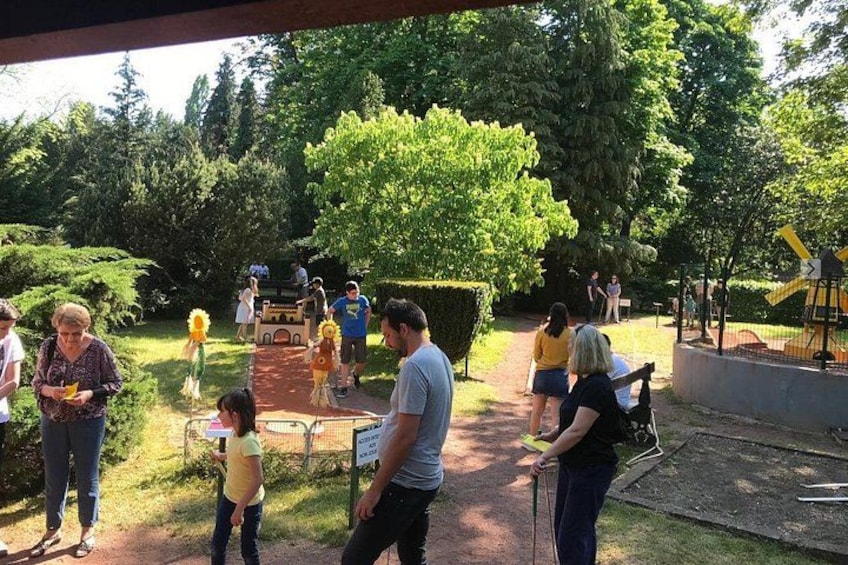 A group of people in a park with a playground, trees, and a paved path. 1