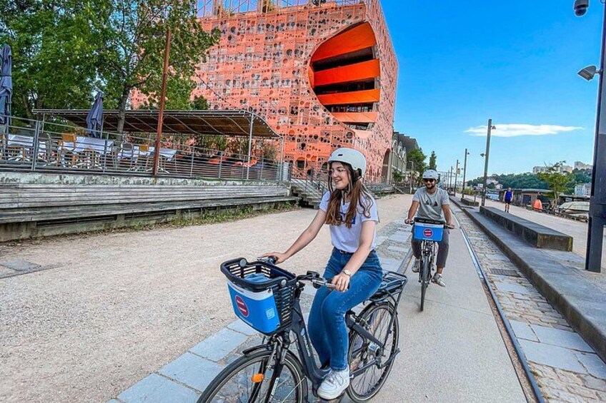 Two people riding bicycles on a paved path with a modern building in the background. 2