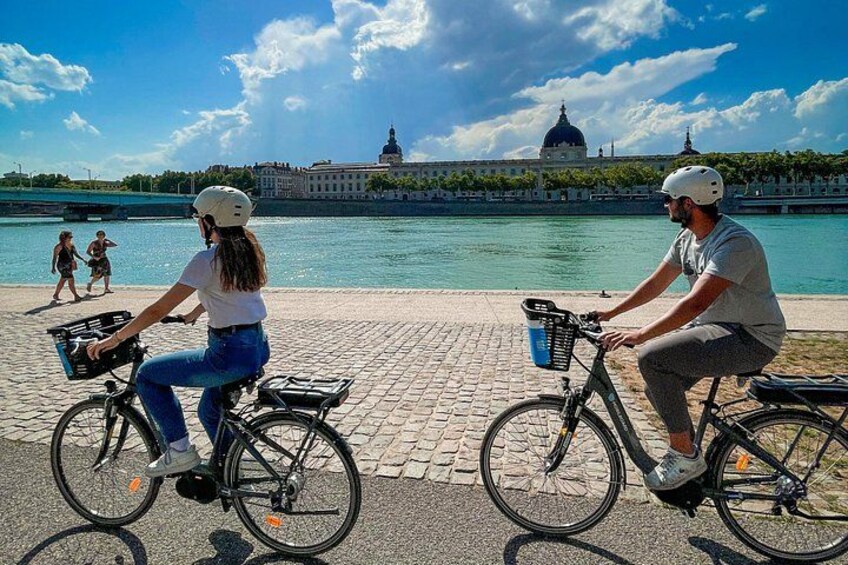 Two people on bicycles with baskets, a river, and a building with a dome in the background. 1