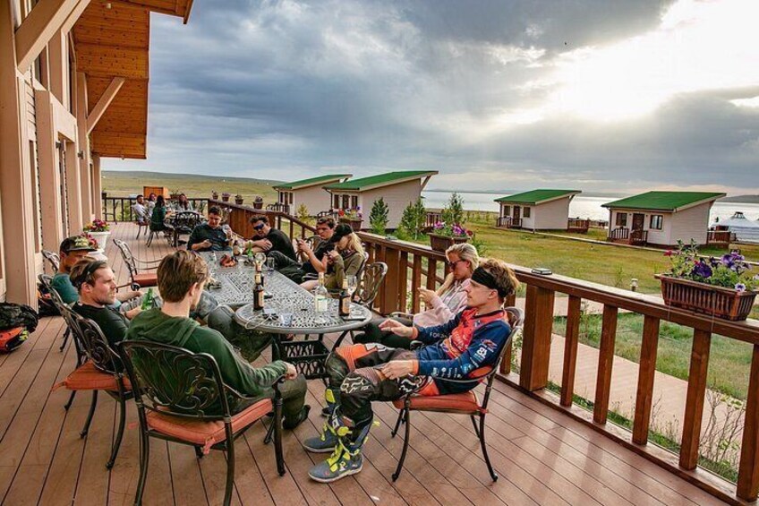 A group of people sitting around a table on a wooden deck with a view of houses and a body of water in the background. 3