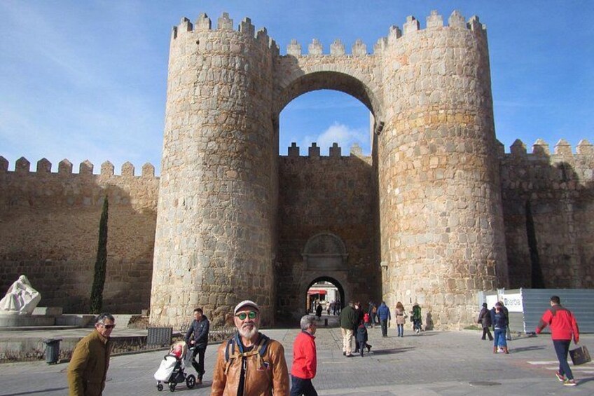 A historic stone archway with battlements, surrounded by a paved area with people walking and a stroller. 2