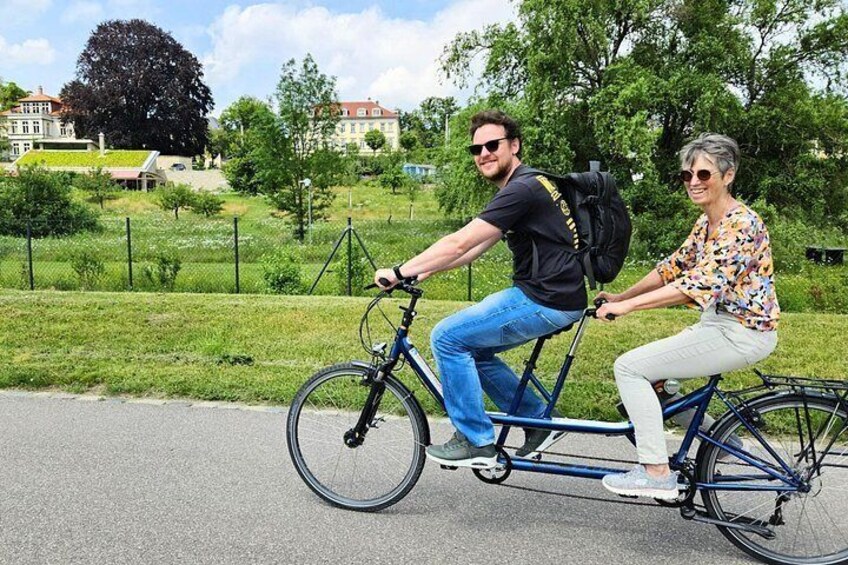 Two people riding a tandem bicycle on a paved path with grass and trees in the background. 3