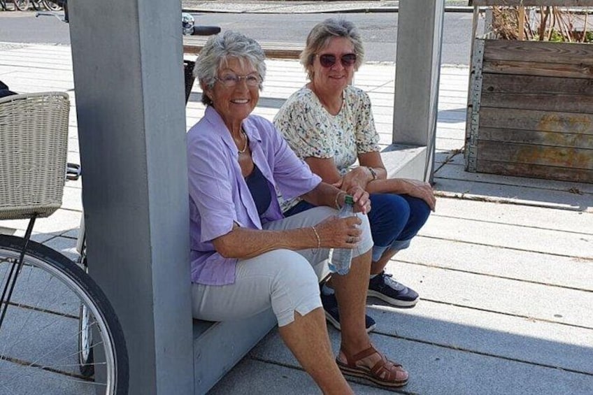 Two people sitting on a bench under a shelter, with a bicycle and a wooden structure in the background. 2