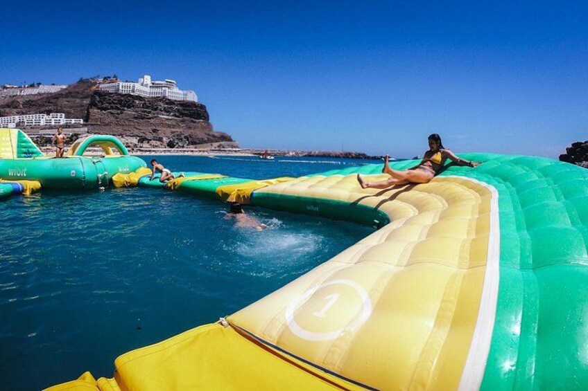A water slide with people using it, a large inflatable structure, and a coastal landscape with a building on a cliff. 2