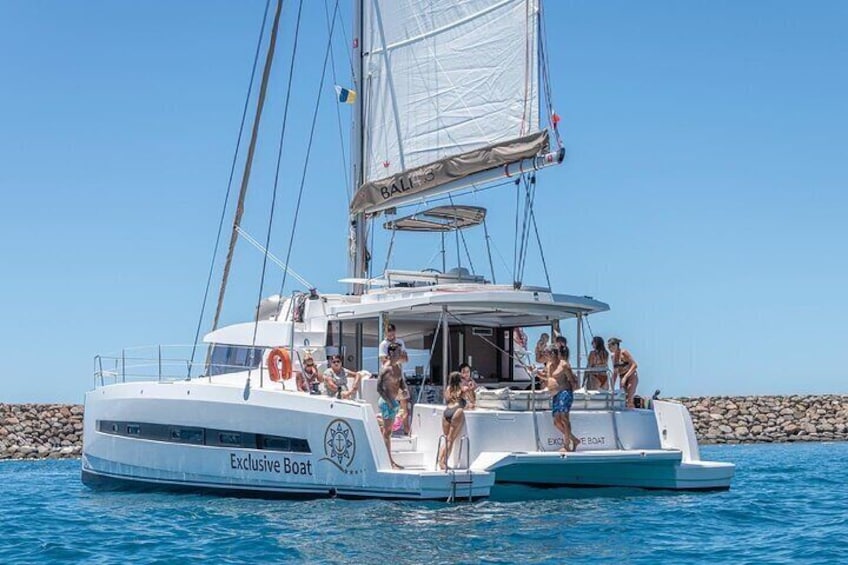 En vit katamaran med namnet ”Exclusive Boat”, med folk ombord, seglar på ett klart, blått hav. 1