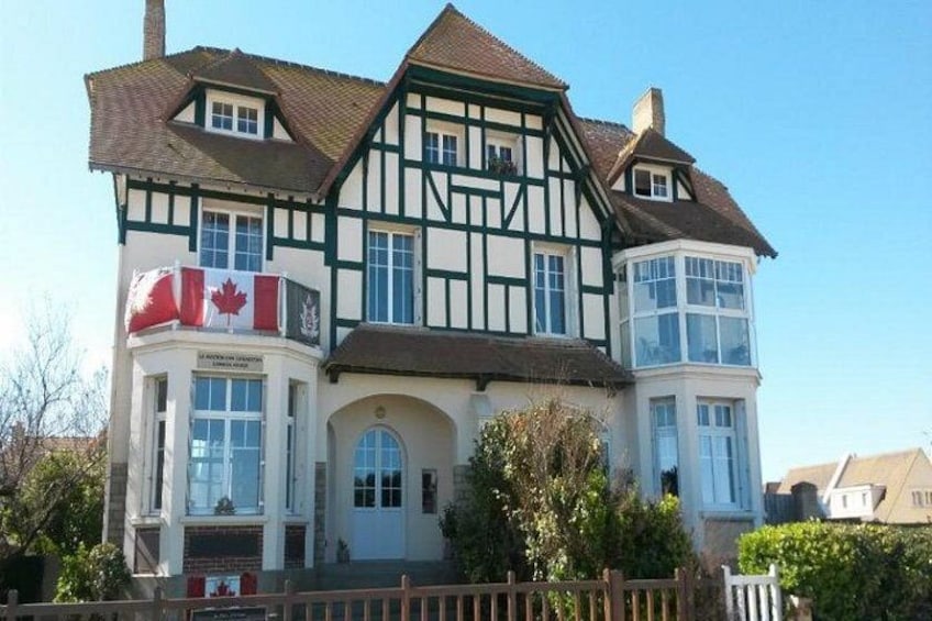 A two-story Tudor-style building with a Canadian flag, a large arched entrance, and a white picket fence. 1